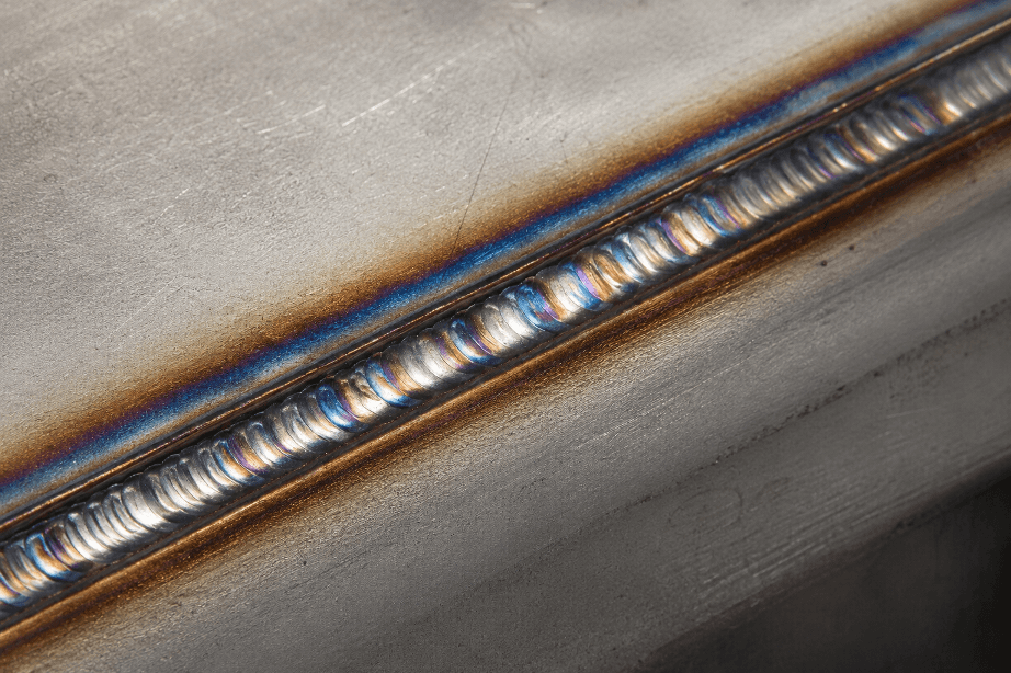 Close-up photograph of a TIG weld showing blue and brown heat tint on stainless steel before cleaning.